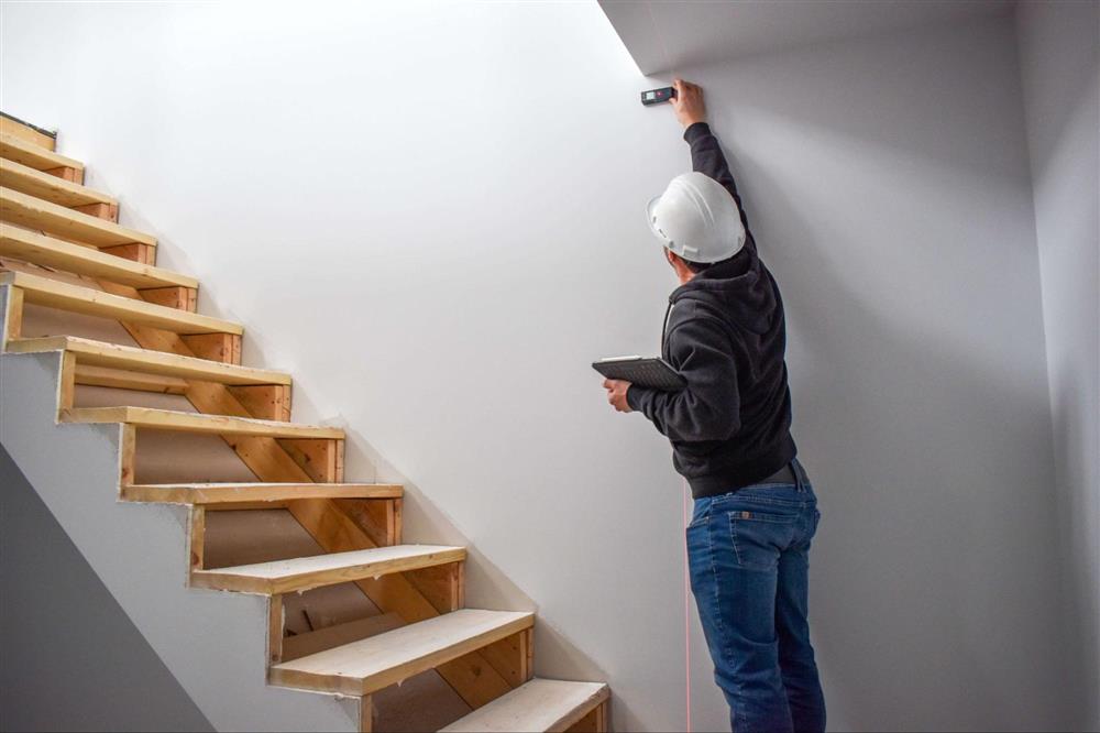 Installateur d’Intermat effectuant des prises de mesures à proximité d’un escalier en bois sur un chantier résidentiel afin d’assurer la coordination des travaux de finition intérieure.